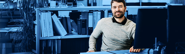A smiling man with a beard sits at a desk in an office, with bookshelves and monitors in the background.